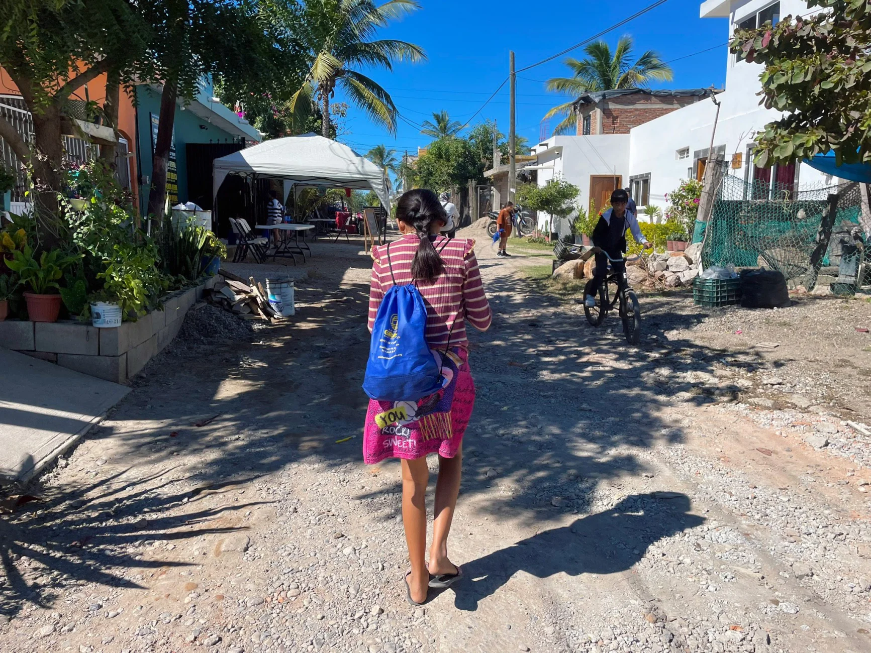 Girl carrying a blue backpack walks down a dirt street in a Mexican coastal village